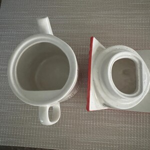 K&ouml;nnte beinhalten: Wei&szlig;e Keramik-Teekanne mit Henkel und Ausguss sowie einem separaten Deckel. Der Deckel hat einen roten Rand. Die Teekanne steht auf einer grauen Tischmatte. Die Teekanne ist zum Aufbr&uuml;hen von Tee bestimmt.