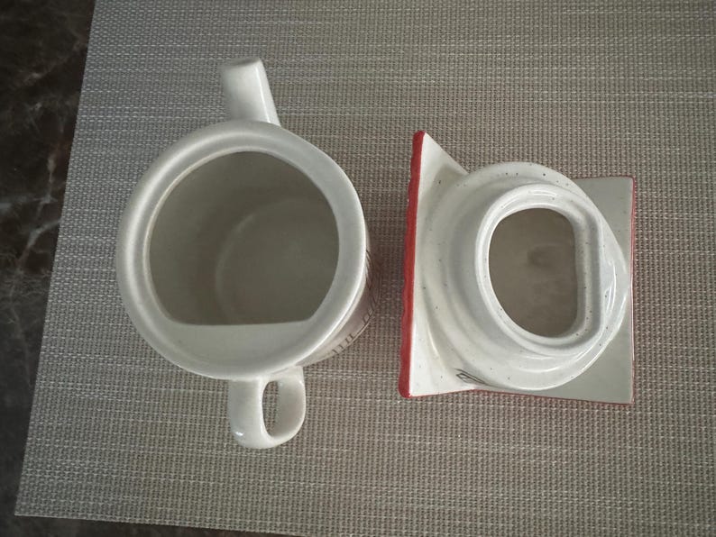 K&ouml;nnte beinhalten: Wei&szlig;e Keramik-Teekanne mit Henkel und Ausguss sowie einem separaten Deckel. Der Deckel hat einen roten Rand. Die Teekanne steht auf einer grauen Tischmatte. Die Teekanne ist zum Aufbr&uuml;hen von Tee bestimmt.