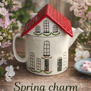 K&ouml;nnte beinhalten: Eine wei&szlig;e Keramik-Teekanne in Form eines Hauses mit rotem Dach. Die Teekanne hat schwarze Fenster- und T&uuml;rdetails und den Text "Spring charm for teatime" auf der Vorderseite. Der Hintergrund zeigt Blumen.