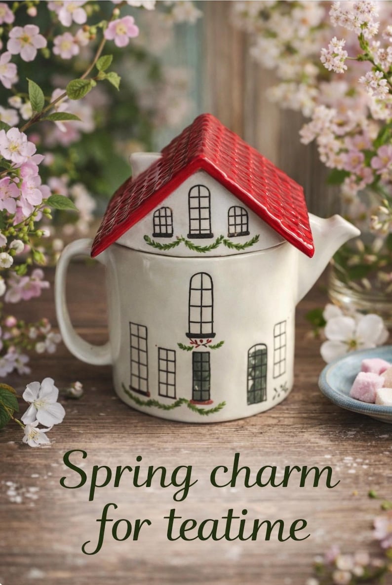 K&ouml;nnte beinhalten: Eine wei&szlig;e Keramik-Teekanne in Form eines Hauses mit rotem Dach. Die Teekanne hat schwarze Fenster- und T&uuml;rdetails und den Text "Spring charm for teatime" auf der Vorderseite. Der Hintergrund zeigt Blumen.