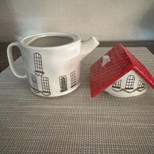 K&ouml;nnte beinhalten: Eine wei&szlig;e Keramik-Teekanne und eine zuckerstreuer in Hausform. Die Teekanne hat schwarze Fensterillustrationen und gr&uuml;ne Girlanden. Der Zuckerstreuer hat ein rotes Dach und &auml;hnliche Fensterdesigns.