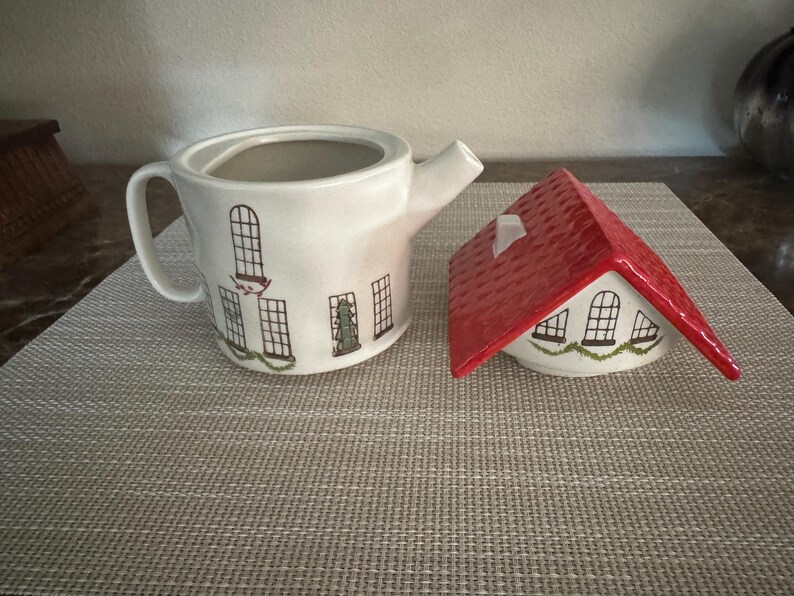K&ouml;nnte beinhalten: Eine wei&szlig;e Keramik-Teekanne und eine zuckerstreuer in Hausform. Die Teekanne hat schwarze Fensterillustrationen und gr&uuml;ne Girlanden. Der Zuckerstreuer hat ein rotes Dach und &auml;hnliche Fensterdesigns.