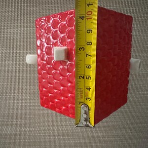 K&ouml;nnte beinhalten: Ein rotes, strukturiertes, rechteckiges Objekt mit wei&szlig;en zylindrischen und quadratischen Akzenten. Das Objekt ist etwa 10 cm hoch, gemessen mit einem gelben Ma&szlig;band. Das Objekt hat ein Muster aus kleinen, erhabenen Kreisen.