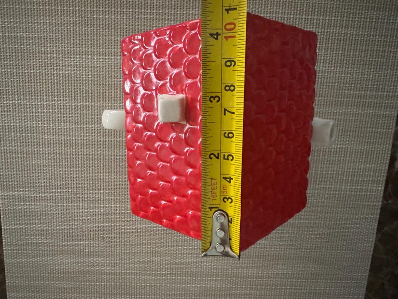 K&ouml;nnte beinhalten: Ein rotes, strukturiertes, rechteckiges Objekt mit wei&szlig;en zylindrischen und quadratischen Akzenten. Das Objekt ist etwa 10 cm hoch, gemessen mit einem gelben Ma&szlig;band. Das Objekt hat ein Muster aus kleinen, erhabenen Kreisen.