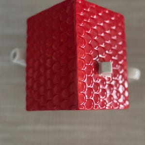 K&ouml;nnte beinhalten: Roter, w&uuml;rfelf&ouml;rmiger Dekorationsgegenstand mit schuppenartiger Textur. Er hat einen kleinen, quadratischen, wei&szlig;en Knopf und zwei wei&szlig;e, zylindrische Griffe. Das Objekt ist wahrscheinlich ein dekoratives Element.