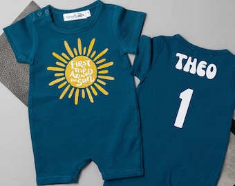 Peut inclure: Deux barboteuses de bébé de couleur teal. L'une présente un soleil jaune avec le texte "First Trip Around the Sun". L'autre porte le nom "THEO" et le chiffre "1" imprimés en blanc au dos.