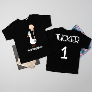 May include: Black t-shirt with a white goose wearing a party hat and holding a balloon. The text "One Silly Goose" is printed below the goose. The back of the shirt has the name "TUCKER" and the number "1" printed in white.