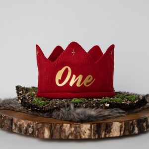 May include: Red knitted crown with gold embroidery that says "One" on a wooden background.
