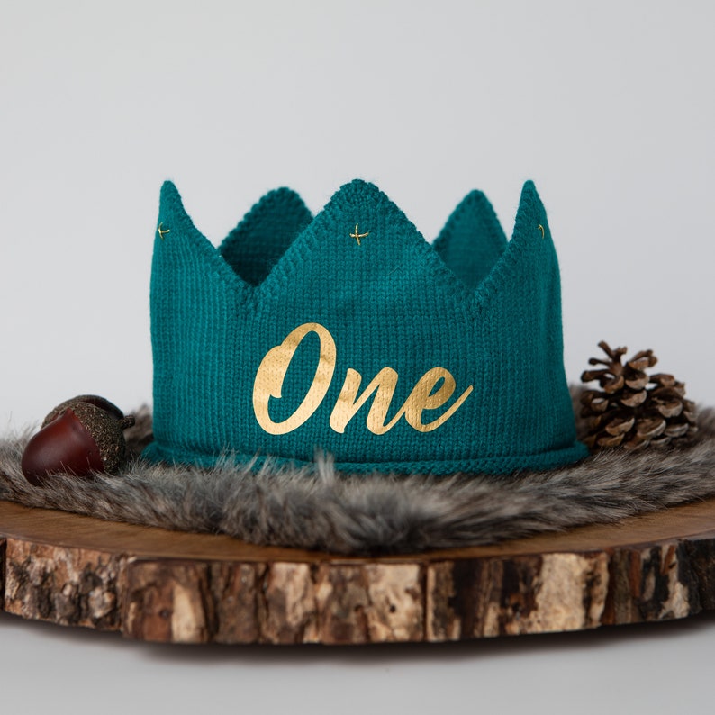 May include: Teal knitted crown with gold lettering that says "One" on a wooden surface with a faux fur trim.