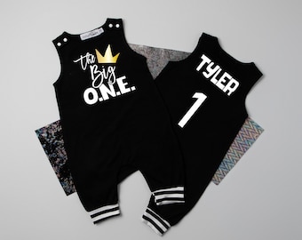 May include: Black sleeveless baby onesie with white text that reads "The Big O.N.E." and a gold crown. The back of the onesie has white text that reads "Tyler 1".