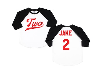 May include: A white and black baseball-style shirt with red text. The front of the shirt says "Two" in a red, stylized font. The back of the shirt says "JAKE 2" in red.