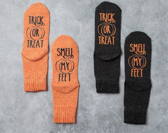 May include: Four pairs of Halloween-themed socks. Two pairs are orange, and two are dark gray. Each sock features a pumpkin graphic and the phrases "TRICK OR TREAT" or "SMELL MY FEET" in black lettering. The socks are made of a textured material.