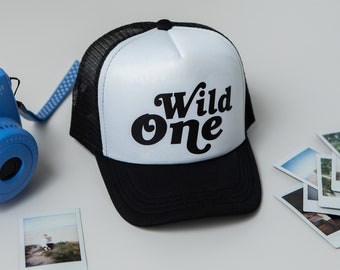 May include: A white and black trucker hat with a black mesh back. The front of the hat has the words "Wild One" in a black retro font.