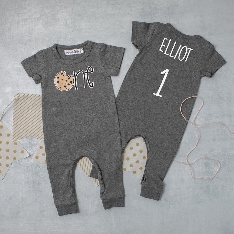 May include: Gray baby onesie with a cookie graphic and the word "one" on the front. The back of the onesie has the name "Elliot" and the number "1" in white.