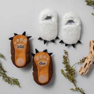 May include: Two pairs of baby booties. One pair is brown with a paw print and the words "Wild One" on them. The other pair is white and fuzzy with black claws.