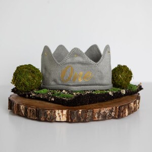 May include: A gray knitted crown with the word "One" in gold lettering on a wooden slice with moss.
