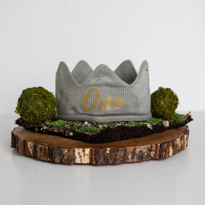 May include: A gray knitted crown with the word "One" in gold lettering on a wooden slice with moss.