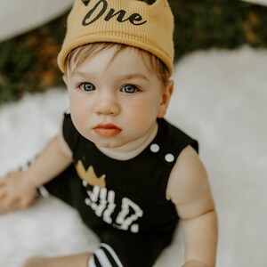 May include: A baby wearing a black and white striped outfit with a gold crown and the word "One" on it. The baby is wearing a yellow knit hat with the word "One" on it. The baby is sitting on a white surface.