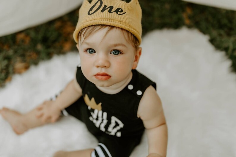 May include: A baby wearing a black and white striped outfit with a gold crown and the word "One" on it. The baby is wearing a yellow knit hat with the word "One" on it. The baby is sitting on a white surface.