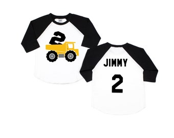 May include: A white and black baseball style shirt with a yellow dump truck carrying the number 2 on the front. The back of the shirt has the name "JIMMY" and the number "2" in black.