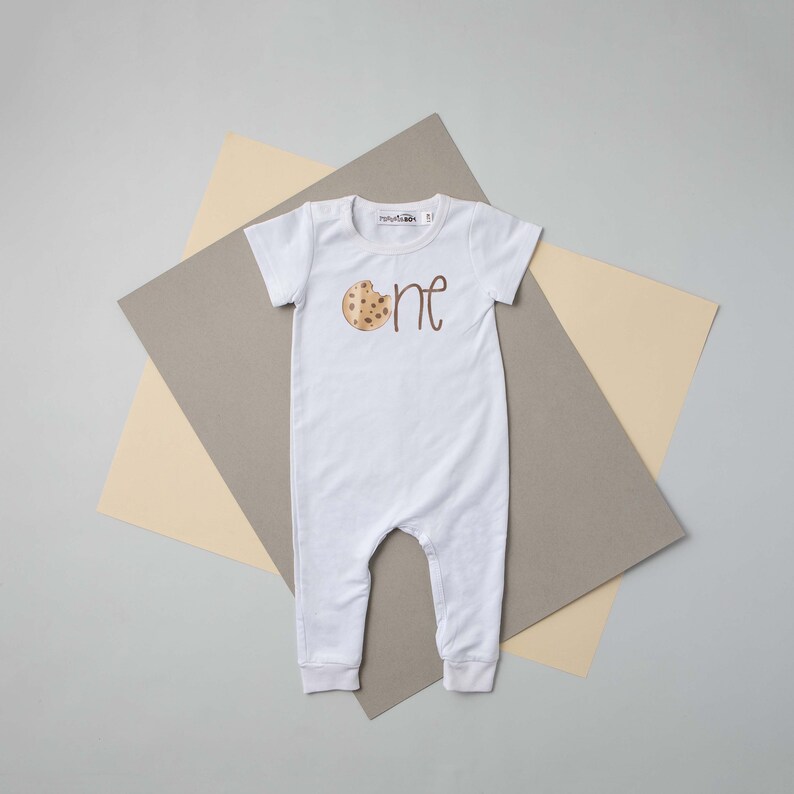 May include: White baby onesie with a brown cookie graphic and the word "one" in brown script.
