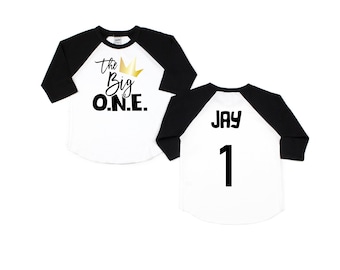 May include: Two white and black baseball shirts with a gold crown and the text "The Big O.N.E." on one shirt and the text "Jay 1" on the other shirt.