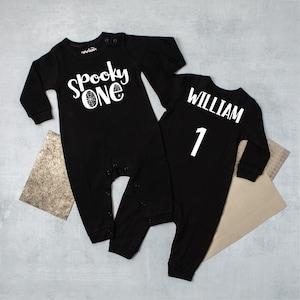May include: Two black baby onesies. One says "Spooky ONE" in white script with a spiderweb graphic. The other says "WILLIAM" and the number "1" in white. Both have long sleeves and snap closures.