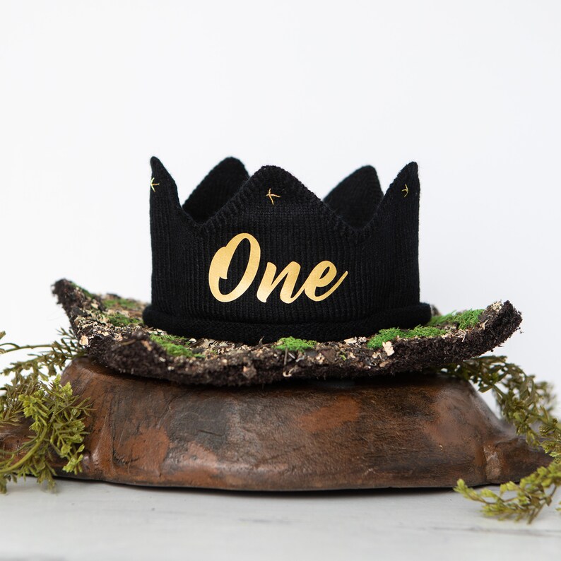 May include: Black knit crown with gold lettering that says "One" on a brown and green mossy base.