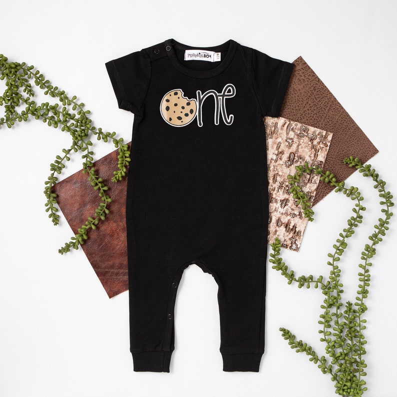 May include: Black short-sleeved onesie with a white graphic of a chocolate chip cookie with a bite taken out of it and the word "one" written in white.