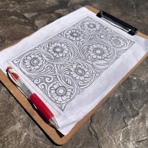 May include: A black and white line drawing of a floral design with sunflowers on a white sheet of paper on a clipboard with a red pen.