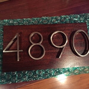 K&ouml;nnte beinhalten: Nahaufnahme einer Hausnummerntafel mit der Nummer 4890 in silbernen Metallbuchstaben auf einem dunkelbraunen Holzuntergrund.