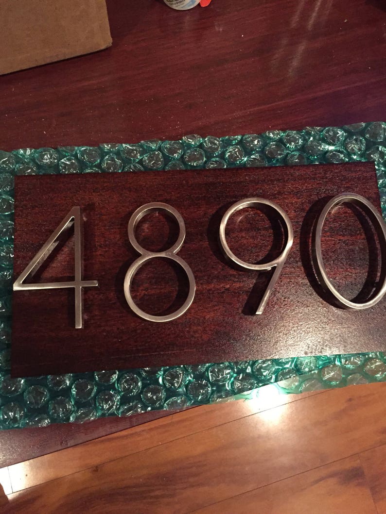 K&ouml;nnte beinhalten: Nahaufnahme einer Hausnummerntafel mit der Nummer 4890 in silbernen Metallbuchstaben auf einem dunkelbraunen Holzuntergrund.