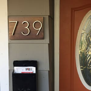 K&ouml;nnte beinhalten: Ein h&ouml;lzernes Hausnummernschild mit der Nummer 739 in goldenen Buchstaben. Das Schild ist an einer hellgrauen Wand neben einer roten T&uuml;r mit einem Glasfenster angebracht.