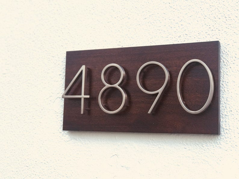 K&ouml;nnte beinhalten: Ein dunkelbraunes Holzschild mit der Nummer 4890 in silbernen Metallbuchstaben.
