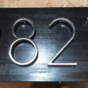 K&ouml;nnte beinhalten: Ein schwarzer Hintergrund mit silbernen Hausnummern aus Metall, die "2827" anzeigen.