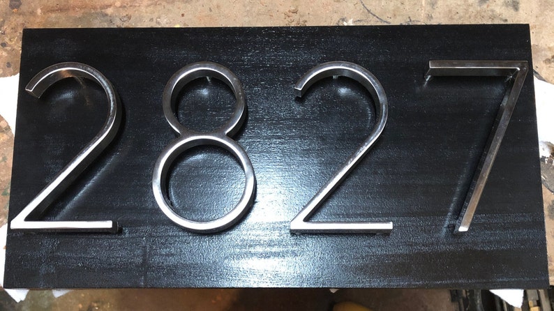 K&ouml;nnte beinhalten: Ein schwarzer Hintergrund mit silbernen Hausnummern aus Metall, die "2827" anzeigen.