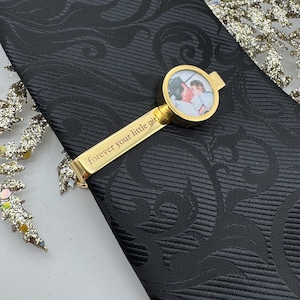 May include: A black necktie with a tonal damask pattern featuring a gold-toned tie bar. The tie bar includes a circular frame containing a small photograph of two people and the engraved text "forever your little girl" along the metal bar.