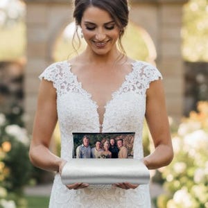 May include: A bride in a white lace wedding dress holds a silver photo box. The dress features a V-neckline and a flowing skirt. The photo box displays a family photo. The background includes a brick pathway and white flowers.