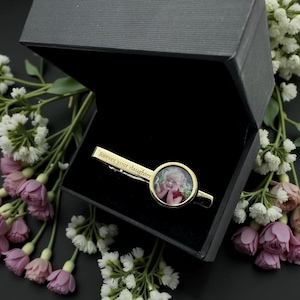 May include: A gold-colored tie clip with a photo of a person inside a circular frame. The clip is engraved with the words "forever your daughter." The tie clip is presented in a black gift box, surrounded by white and pink flowers.