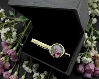 May include: A gold-colored tie clip with a photo of a person inside a circular frame. The clip is engraved with the words "forever your daughter." The tie clip is presented in a black gift box, surrounded by white and pink flowers.