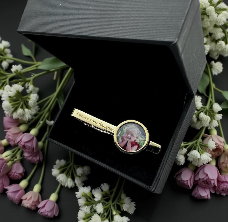 May include: A gold-toned tie clip featuring a circular photo insert of a smiling child and the engraved text "forever your daughter" on the bar, presented inside a black hinged gift box with a velvet-lined interior and a matte finish.