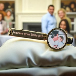 May include: A gold-toned metal tie clip featuring a circular frame containing a photograph of an adult kissing a child on the cheek. The rectangular bar of the tie clip is engraved with the text "forever your little girl" in a serif font.