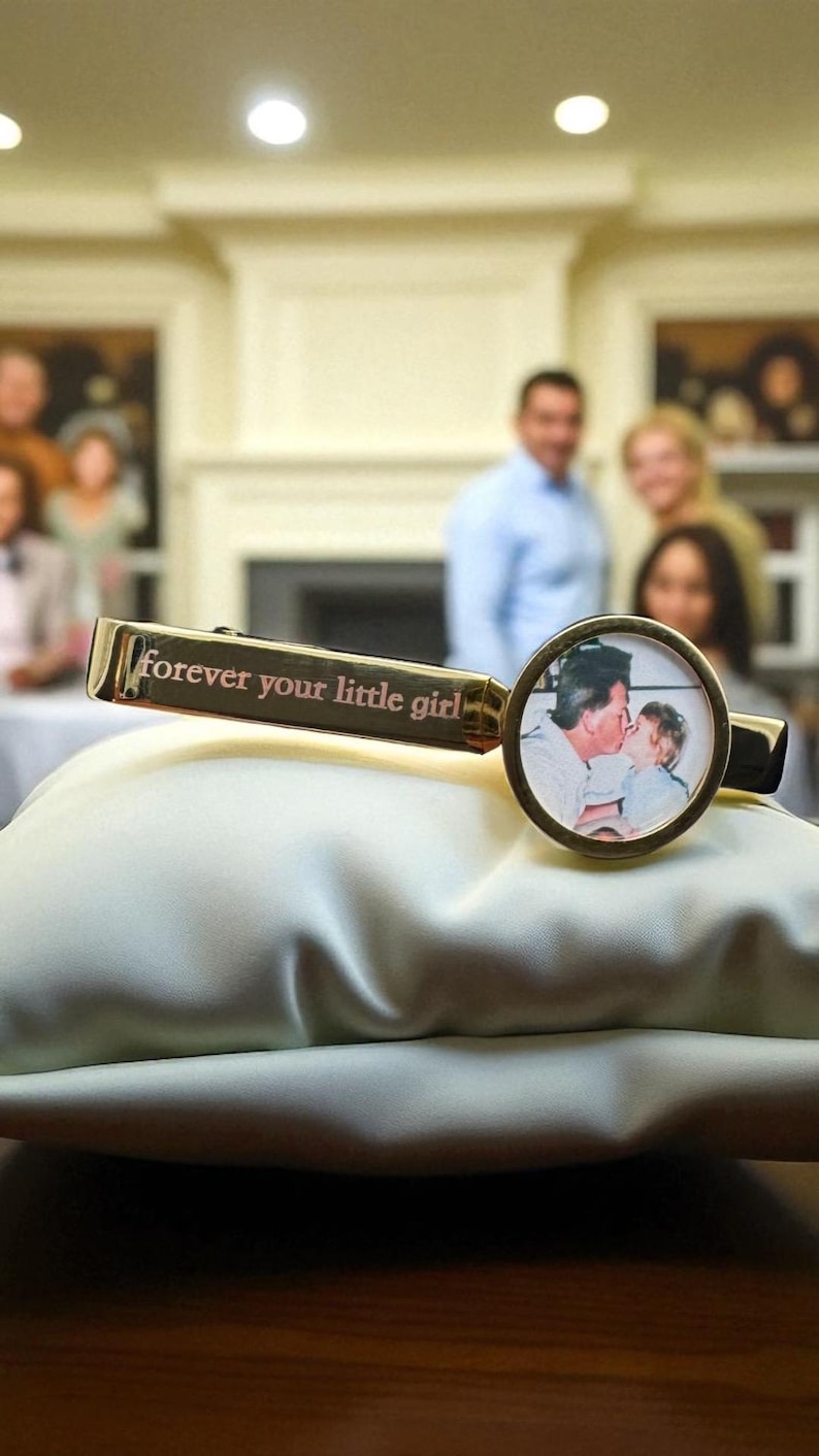 May include: A gold-toned metal tie clip featuring a circular frame containing a photograph of an adult kissing a child on the cheek. The rectangular bar of the tie clip is engraved with the text "forever your little girl" in a serif font.
