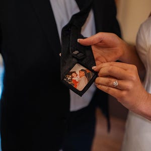 Pode incluir: Uma gravata preta com uma pequena foto de um homem e uma criança em um vestido vermelho. A foto é emoldurada por uma borda preta.