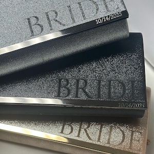 May include: Three glittery clutch purses in silver, black, and rose gold, each with a metallic trim. The word "BRIDE" is engraved on each purse, along with the date "10/14/2025".