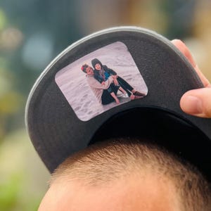 May include: A black baseball cap with a personalized photo patch featuring two people sitting on a beach. The photo is in color and shows a man and a woman.