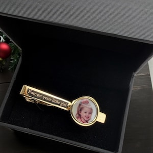 May include: A gold-toned tie clip featuring a circular frame containing a photograph of a young child. The rectangular bar of the tie clip is engraved with the text "Forever your little girl" in a serif font, presented inside a dark gray box.