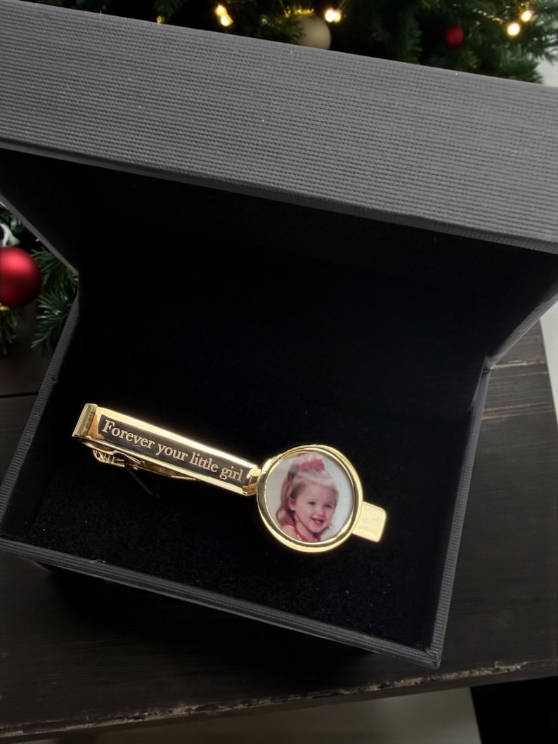 May include: A gold-toned tie clip featuring a circular frame containing a photograph of a young child. The rectangular bar of the tie clip is engraved with the text "Forever your little girl" in a serif font, presented inside a dark gray box.