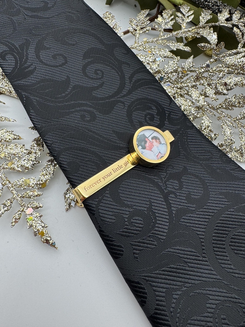 May include: A black necktie with a tonal damask pattern featuring a gold-toned tie bar. The tie bar includes a circular frame containing a small photograph of two people and the engraved text "forever your little girl" along the metal bar.