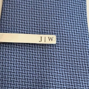 May include: A silver-toned metal tie bar featuring the engraved initials J | W, clipped onto a blue necktie with a repeating geometric herringbone pattern. The tie bar has a smooth, polished finish and is positioned horizontally across the fabric.